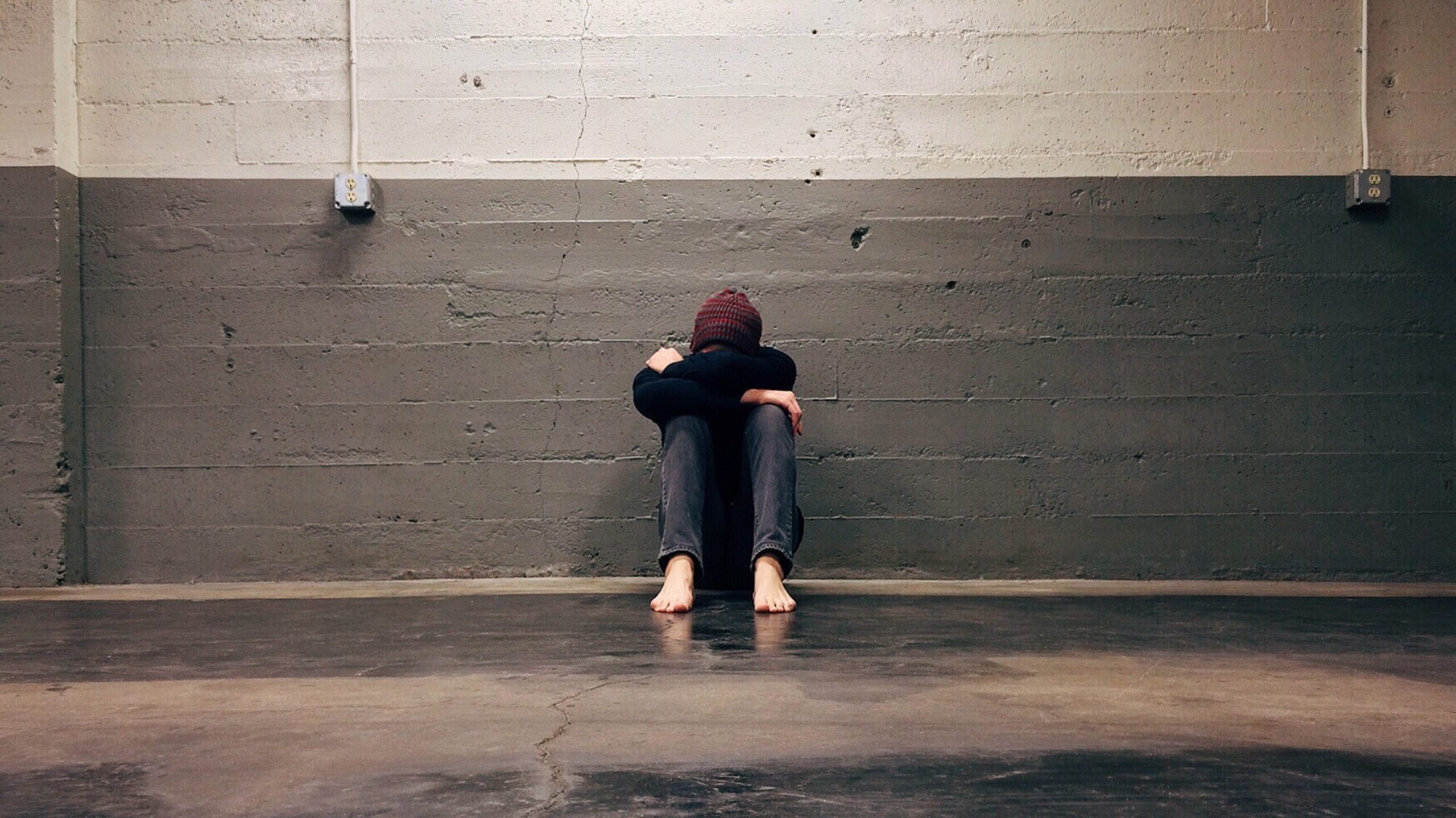 Person sitting on floor against a wall with their head tucked to their knees